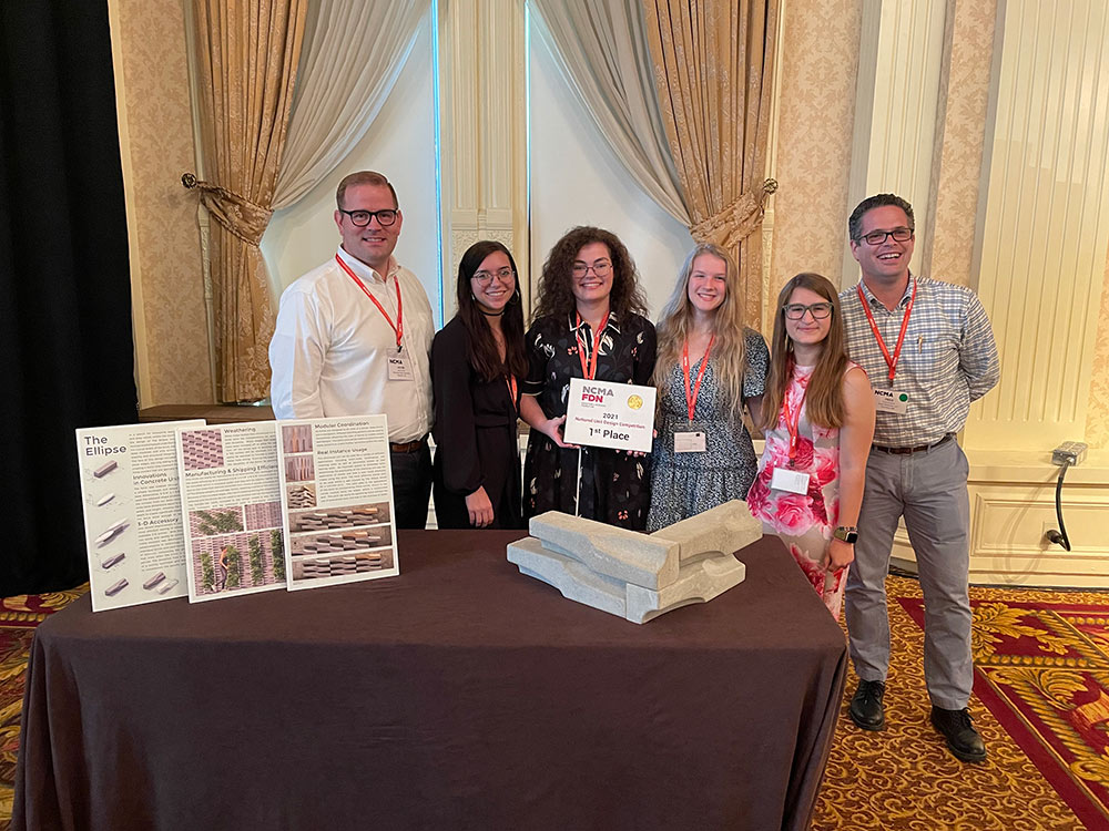 Posing for a photo with first place sign behind table with stacked bricks and presentation photo: (left to right) MSU Associate Professor of Architecture Jacob Gines; MSU architecture juniors Skylar N. Sloan of Saltillo; Sarah E. Mixon of Huntsville, Alabama; Jennifer K. “Jenny” Hutton of Southaven; and Gabriella B. “Gabbi” Morelli of Delmar, Delaware; and Fred Dunand, president and owner of Saturn Materials LLC of Columbus, sponsor of the local Unit Design Competition.